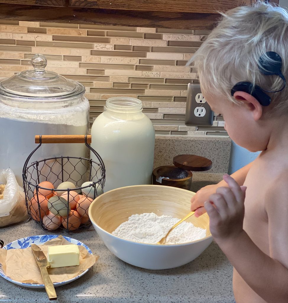 Boy mixing brown sugar pancake mix with a gold fork, eggs are sitting on a counter top in a basket, as well as whole milk and flour