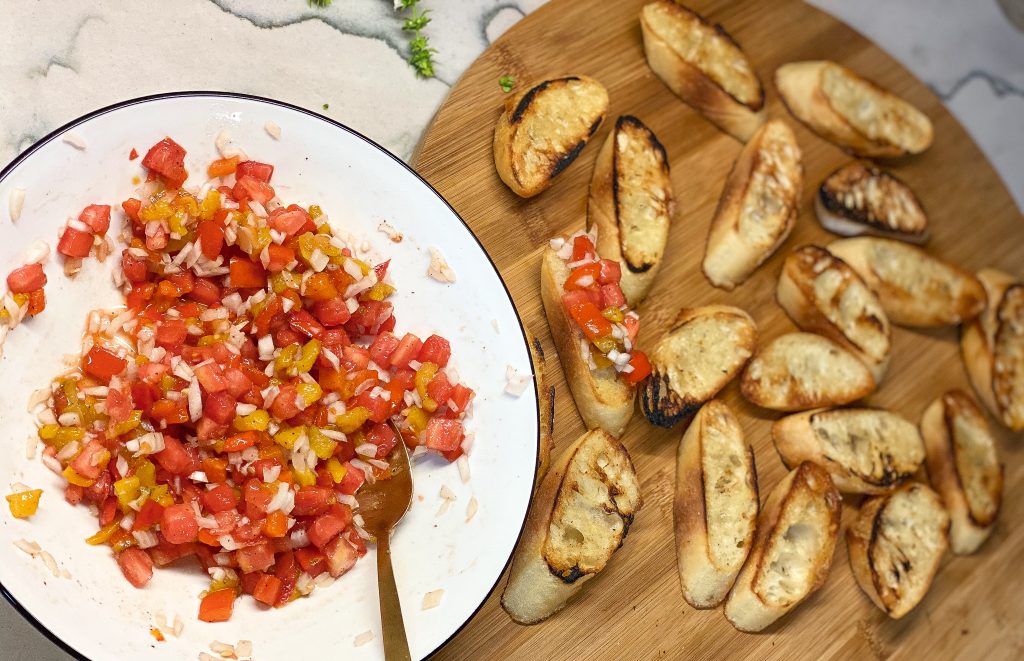 chopped tomato and bell peppers on a white bowl, with a cutting board next to it. with toasted baguette slices.