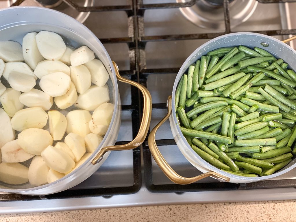 red potatoes cooking in a white pot with golden handles. next to it are green beans cooking in a pot