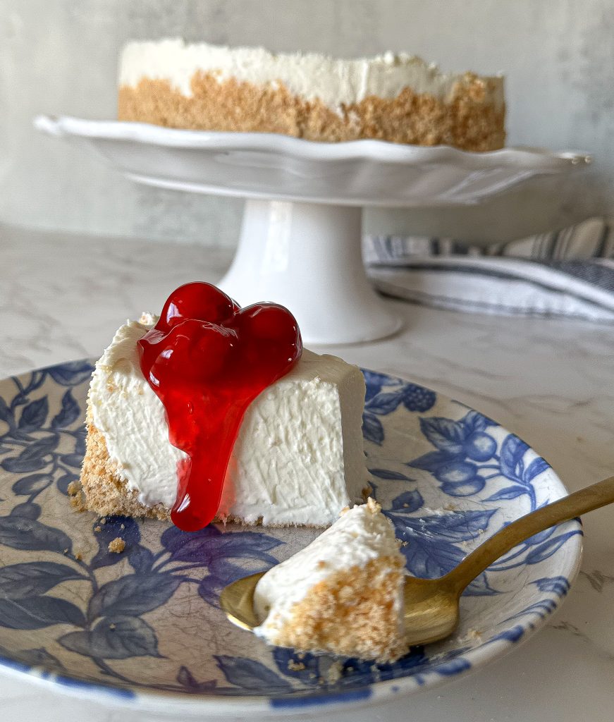 a slice of amazing no bake cheesecake on a white and blue plate with cherry pie filling on top, with a gold spoon