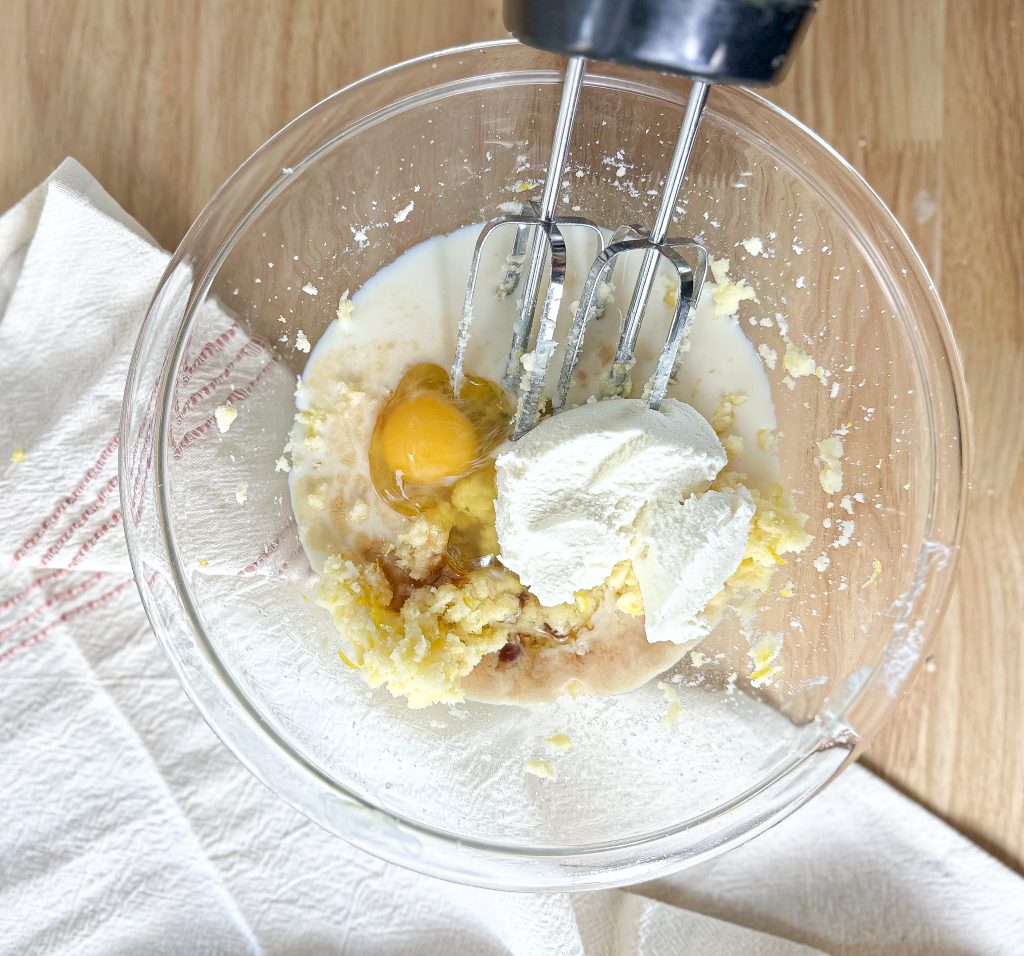 lemon, lemon zest, ricotta cheese, sugar, butter, and egg in a clear bowl with an electric mixer standing on a wooden countertop