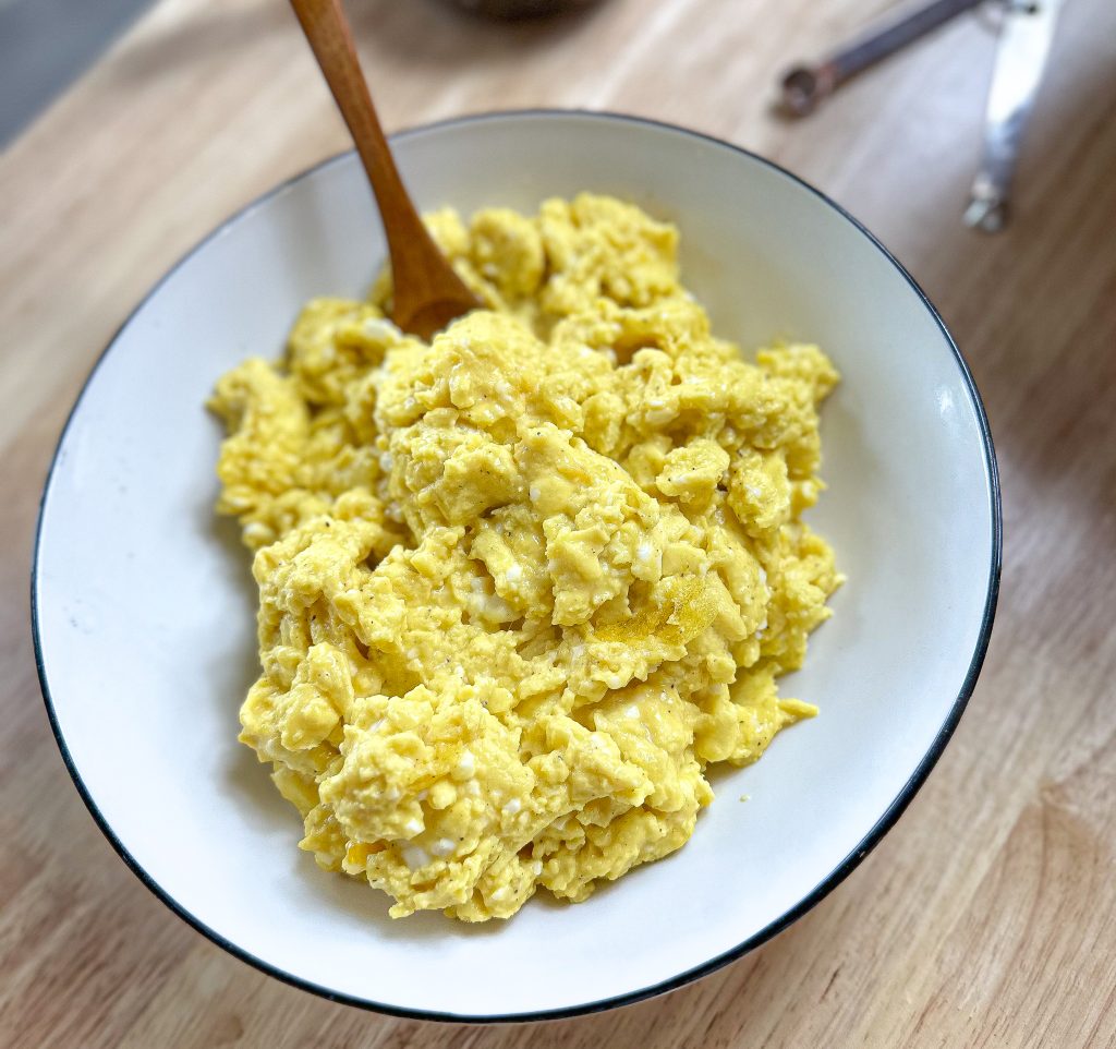 fluffy scrambled eggs in a white bowl with a wooded spoon on butcher block countertop
