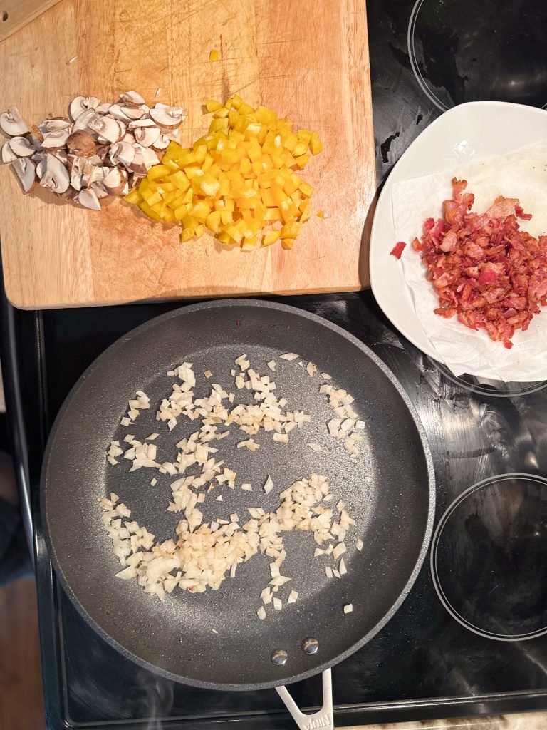 onions sauteing in a skillet on the stove with chopped mushrooms and yellow bell pepper next to it on a butcher block