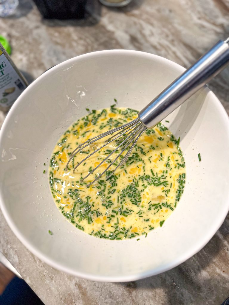 egg mixture with fresh chives in a white mixing bowl and a whisk inside it