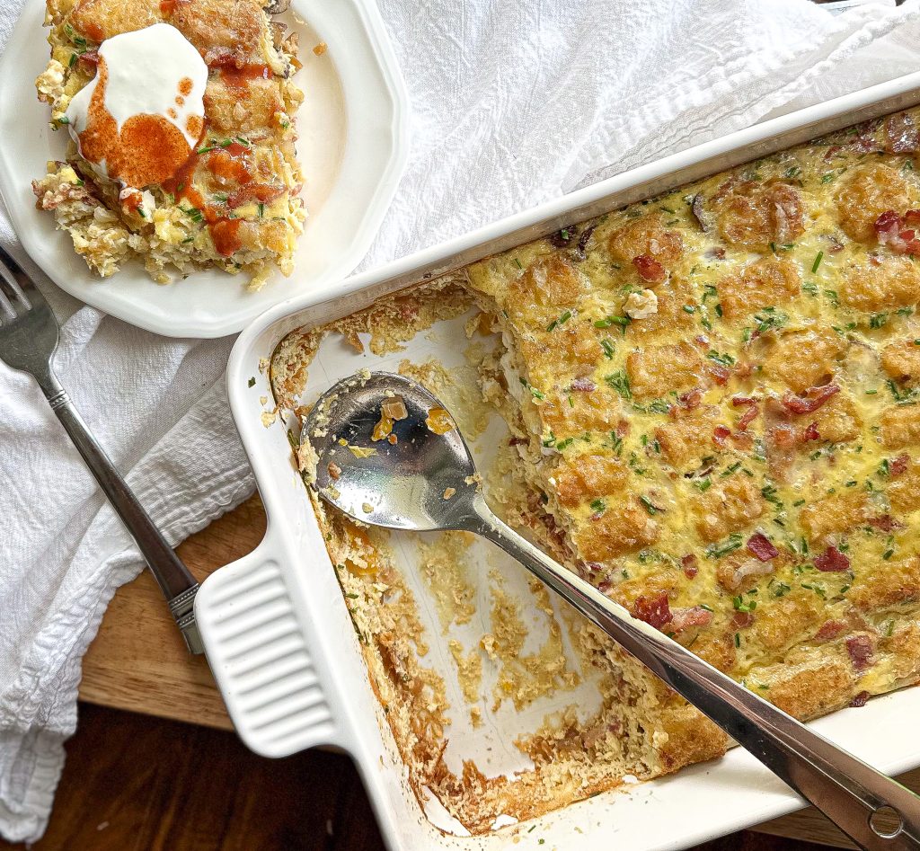 Bacon And Egg Tater Tot Breakfast Casserole with a silver serving spoon in the dish and a serving of the casserole in a small plate with sour cream and hot sauce