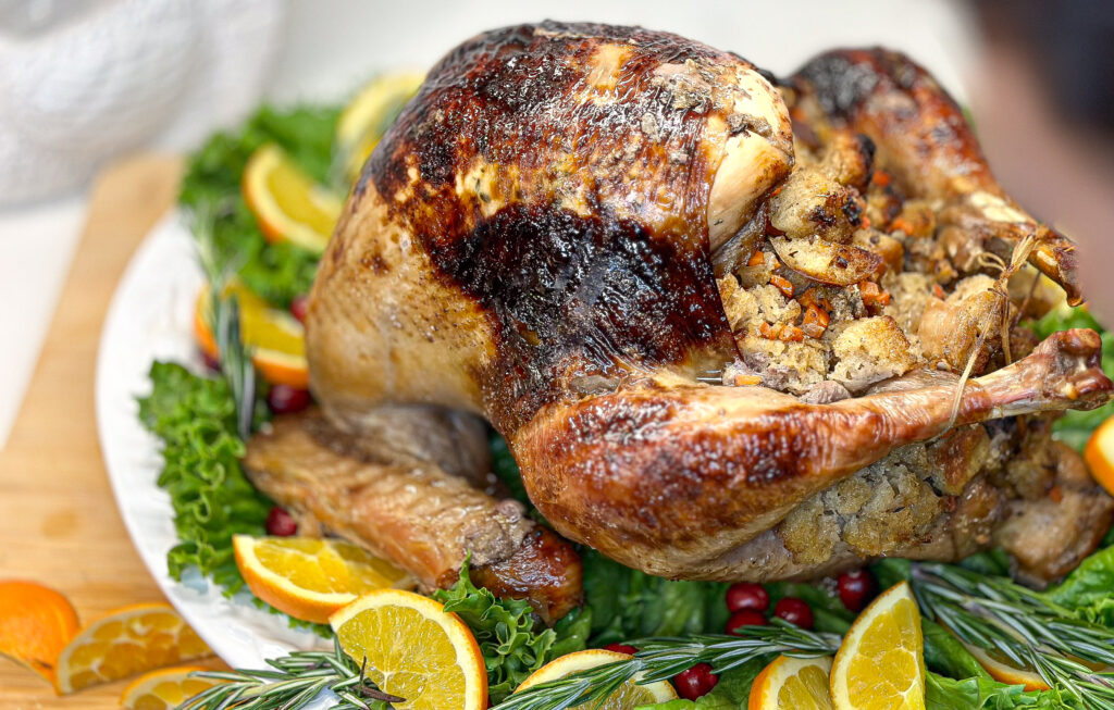 roasted turkey stuffed with homemade sourdough stuffing, the turkey is displayed on a large serving plate with green lettuce underneath the turkey and sliced oranges, rosemary, and cranberries laid around it. 