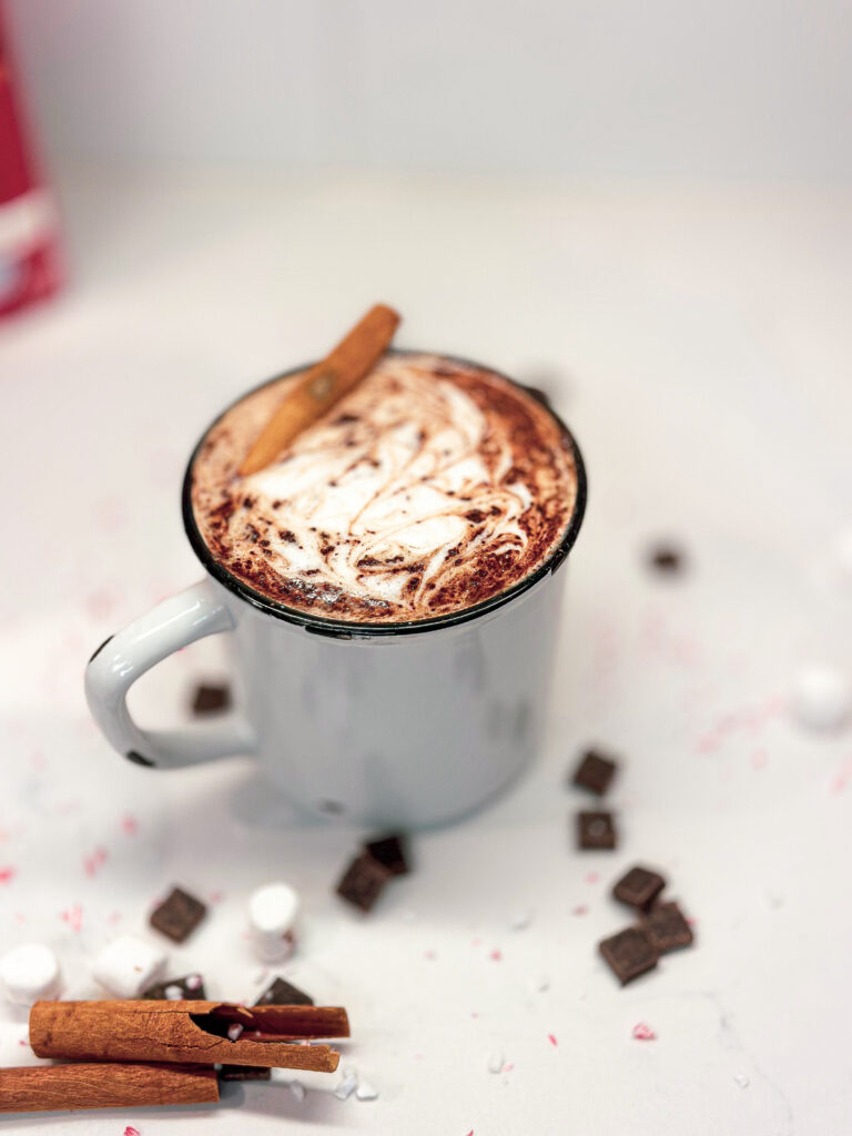 bone broth hot cocoa in a rustic white mug with a cinnamon stick on the inside of the mug. Two cinnamon sticks on a white quartz countertop along with scattered marshmallows, dark chocolate chips and crushed candy canes. 