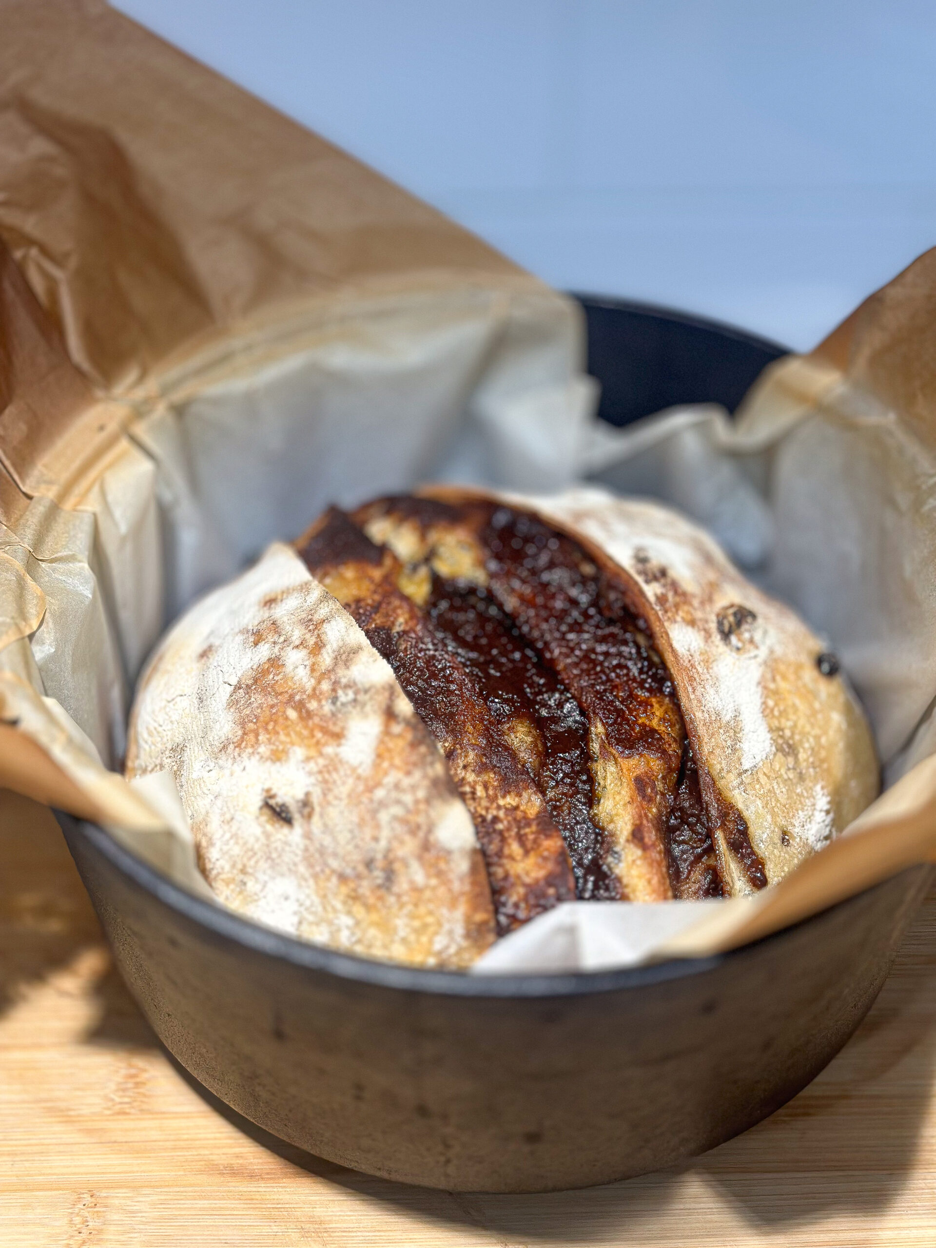 A round loaf of rustic bread with a dark, crusty split on top sits in a parchment-lined black pot on a wooden surface.