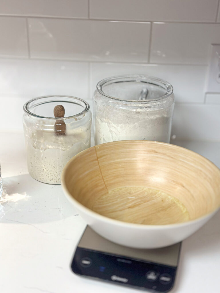 wooden bowl on a food scale with big clear jars behind it with sourdough starter and flour on white  quarts countertop