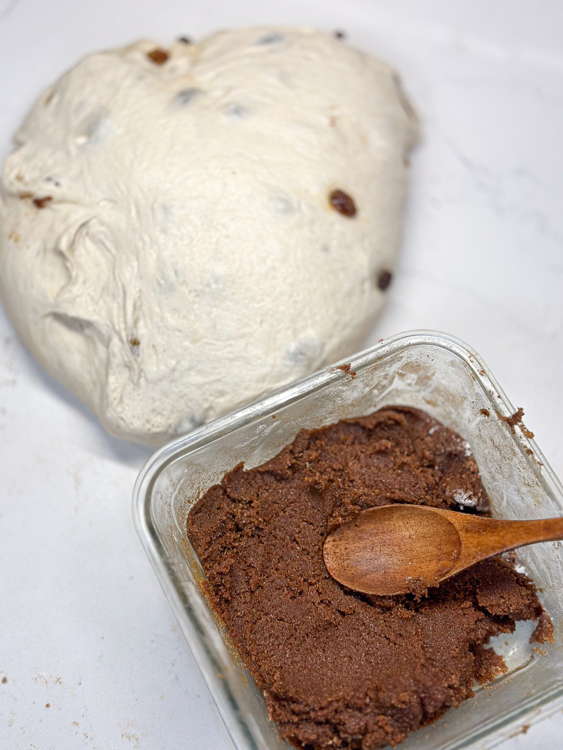 A ball of bread dough with raisins next to a glass container of brown cinnamon sugar mixture and a wooden spoon on a white surface.