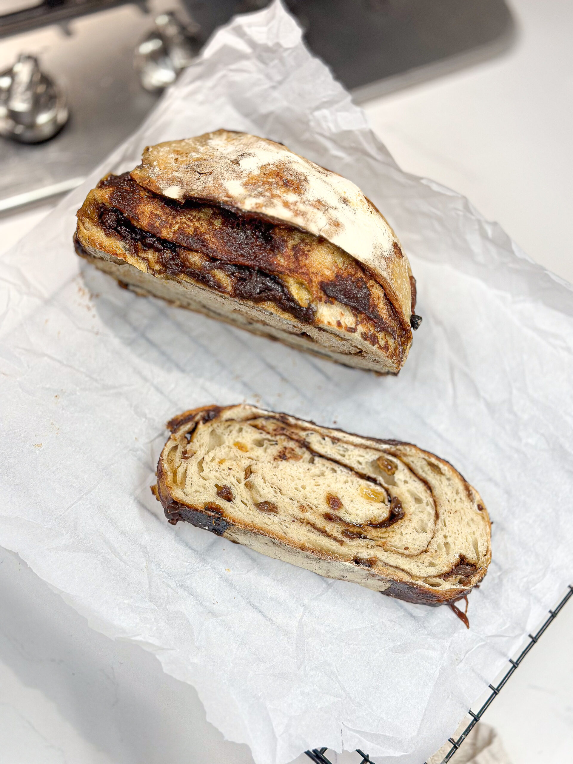 A loaf of cinnamon swirl bread, partially sliced, sits on white parchment paper with one slice placed in front of it.