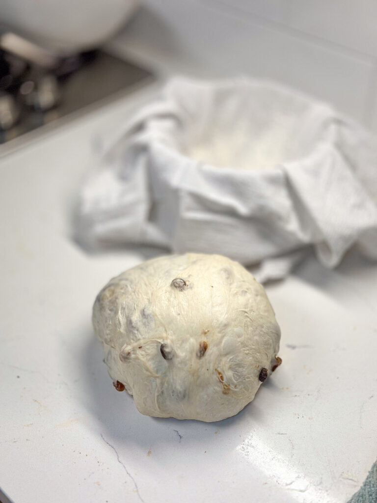 sourdough cinnamon raisin bread shaped into a boule on a white quart countertop with a proofing bowl next to it