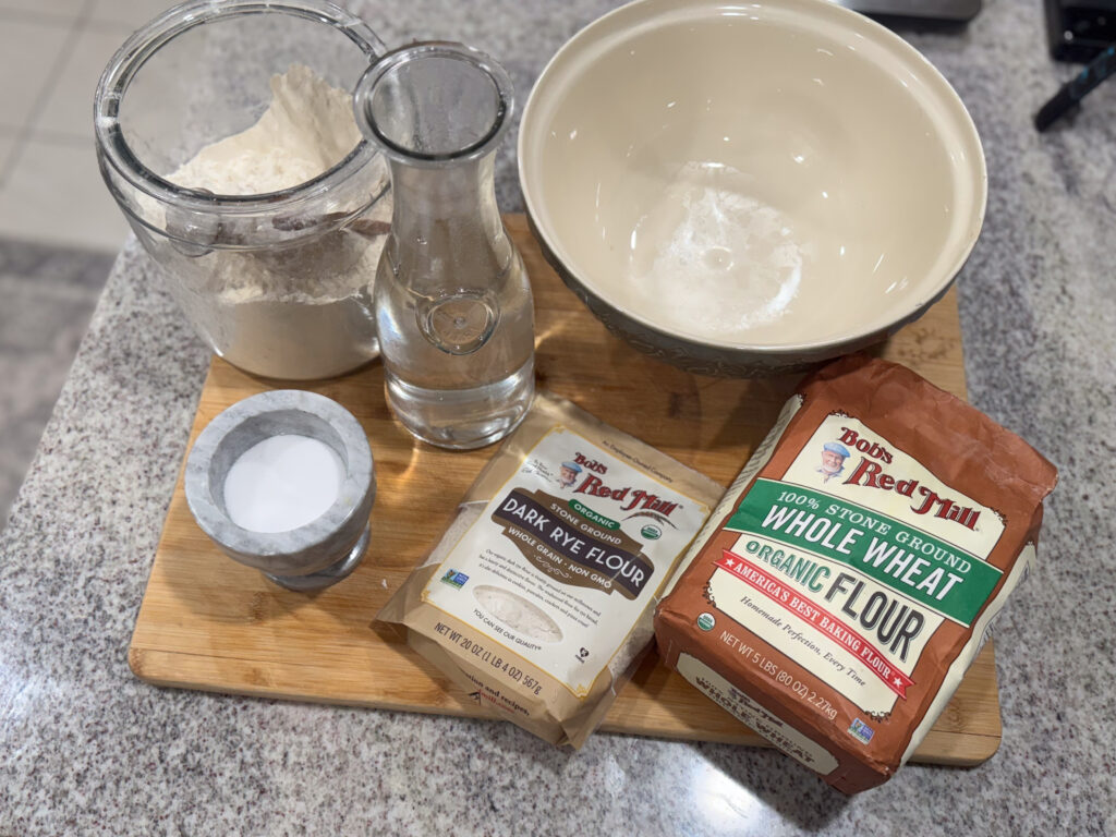 Ingredients for sourdough rye bread arranged on a wooden board, including a jar of flour, a carafe of water, a bowl, a container of salt, Bob’s Red Mill dark rye flour, and whole wheat flour.