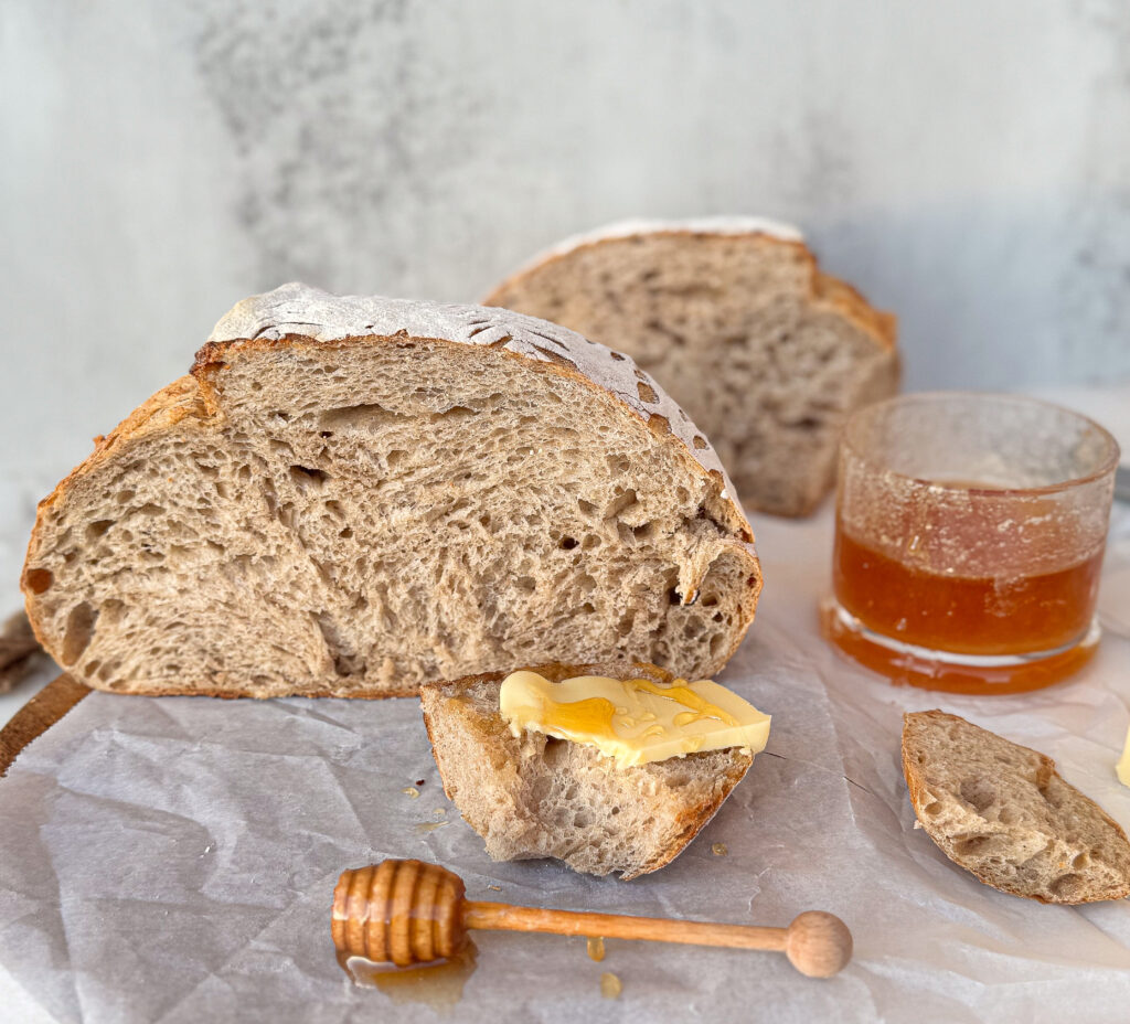 Rustic sourdough rye bread with a crusty exterior and airy crumb, sliced open and served with butter and honey. A glass of honey and a honey dipper rest nearby on parchment paper.