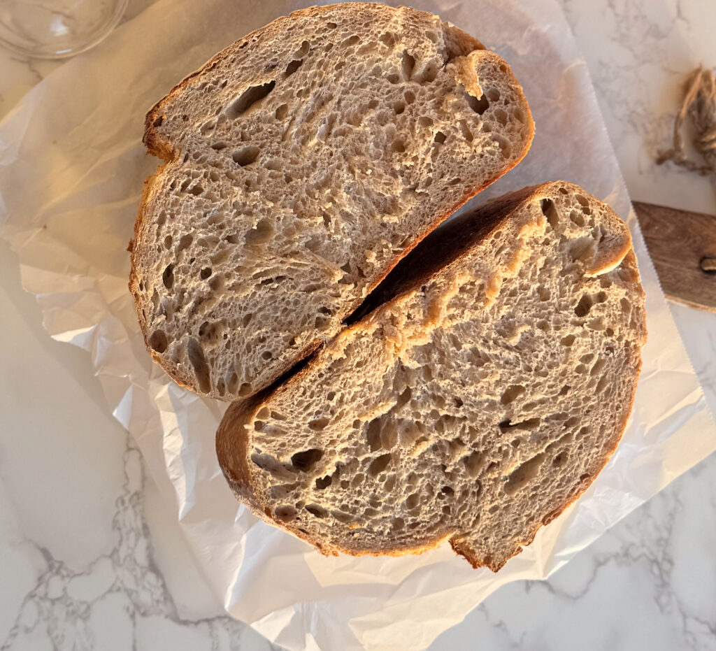 Overhead view of a rustic sourdough rye bread sliced in half, showing its airy crumb and golden crust, placed on crinkled parchment paper over a marble surface.