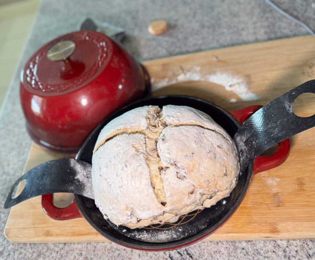 A round loaf of unbaked sourdough s’mores bread sits in a black cast iron pan lined with a floured sling, ready for baking. The dough is dusted with flour and scored with a cross-shaped cut on top. In the background, a red Dutch oven lid rests on a wooden cutting board, with some scattered flour visible on the countertop.