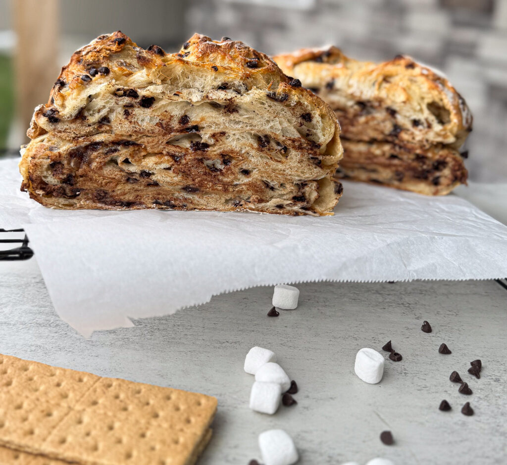 A sliced loaf of sourdough s’mores bread with a golden-brown, crispy crust and a soft, airy interior swirled with melted chocolate, marshmallows, and graham cracker crumbs. The bread sits on a parchment-lined cooling rack, with small marshmallows, chocolate chips, and graham crackers scattered on the light-colored surface nearby. The layers of filling are visible within the bread, creating a marbled effect of chocolate and toasted marshmallow.