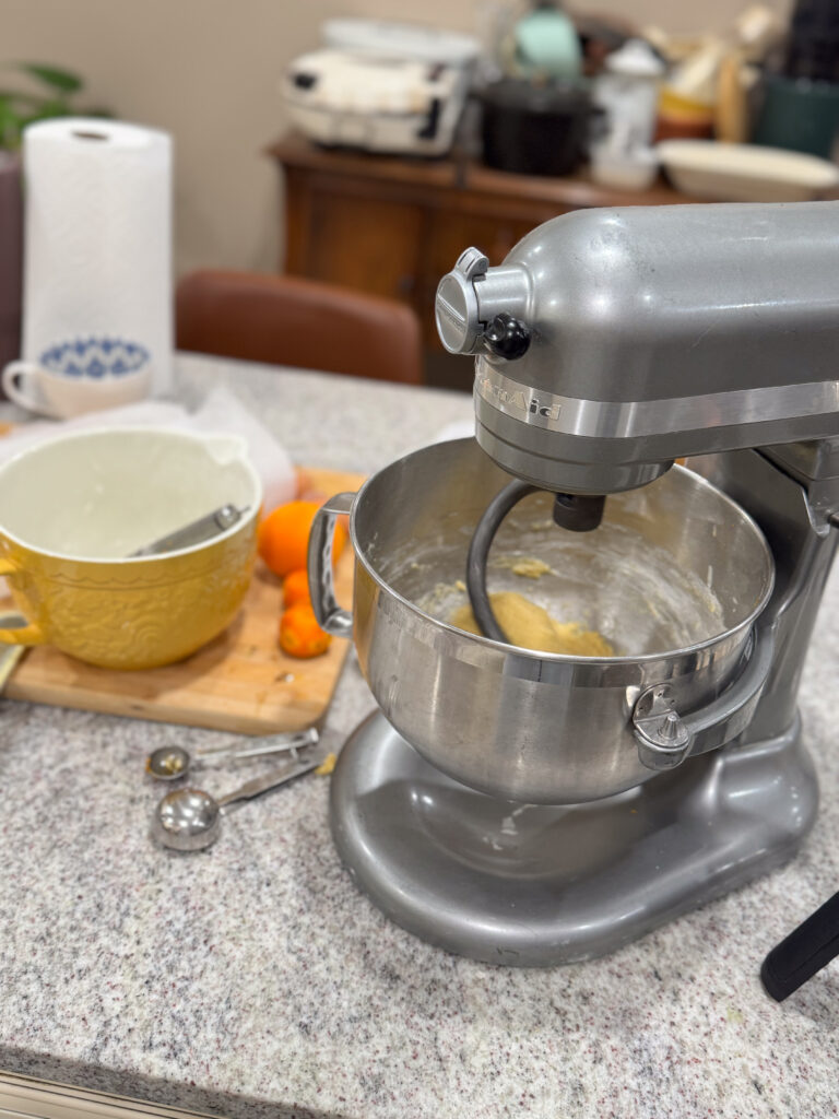 A kitchen countertop with a silver KitchenAid stand mixer in the foreground, mixing dough in a metal bowl. Next to the mixer, there's a yellow ceramic bowl, a cutting board with oranges and measuring spoons scattered around, and a cup in the background. A roll of paper towels is visible on the left, and kitchen items can be seen on the shelves in the background. The scene is bright and cozy, suggesting a homey baking environment.