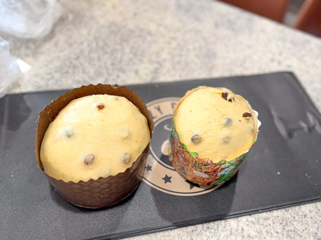 A close-up of two unbaked sourdough Easter bread doughs, each in a different baking paper mold. One is in a dark brown mold, while the other is in a colorful mold with festive designs. Both doughs are slightly risen, showing raisins and orange zest in the mixture. The doughs are placed on a warming mat with a circular logo design visible underneath.