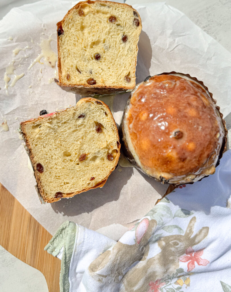 A close-up shot of sliced sourdough Easter bread with raisins, topped with a glossy glaze. The bread has a soft and fluffy crumb, showcasing a light, airy texture with a golden-brown crust. A linen napkin with a bunny and flower design is placed nearby. The scene is bright and sunny, with parchment paper as the backdrop.