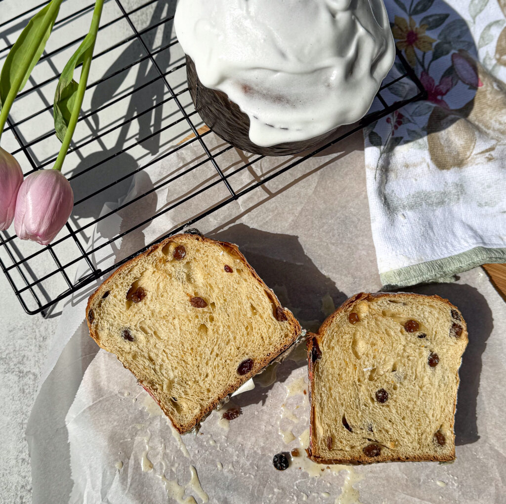 A close-up shot of sliced sourdough Easter bread with raisins, topped with a thick, glossy white glaze. The bread has a soft, fluffy crumb, revealing a light and airy texture with a golden-brown crust. A few pink tulips are placed to the left, and the bread is resting on parchment paper with a linen napkin featuring floral design nearby. A cooling rack is visible in the background with the whole loaf, still adorned with its glaze. The setting is bright, with sunlight highlighting the bread's texture.