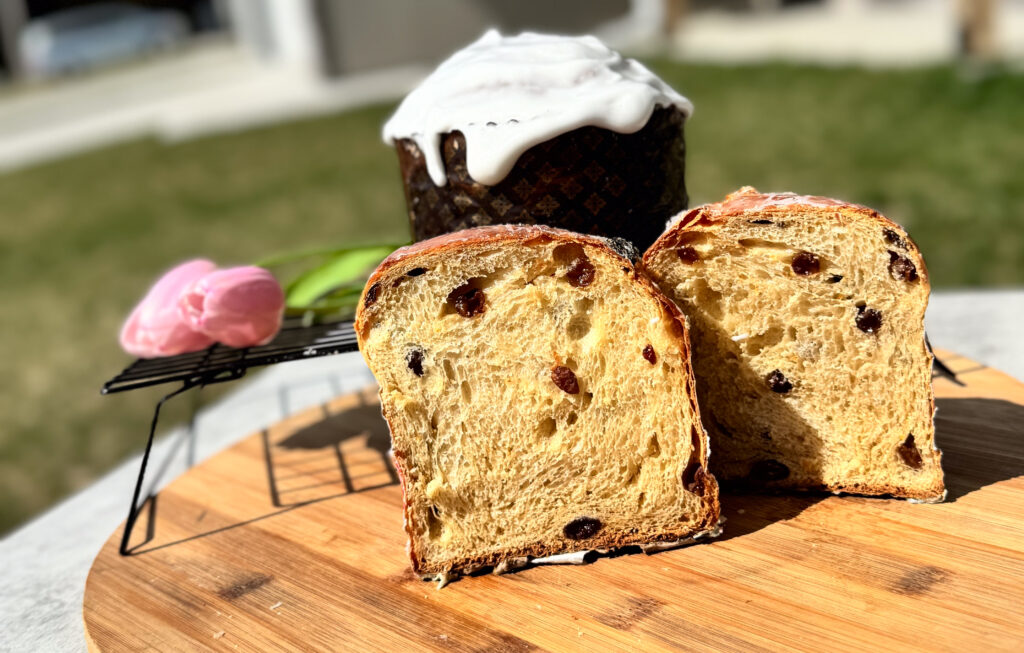 A freshly baked sourdough Easter bread with raisins, sliced to reveal a soft, airy crumb. The bread is placed on a wooden cutting board, with a cooling rack holding the whole loaf in the background. The loaf is topped with a glossy white glaze. A pink tulip is visible in the foreground, adding a touch of spring. The scene is outdoors, with bright sunlight illuminating the bread and creating a warm, inviting atmosphere.