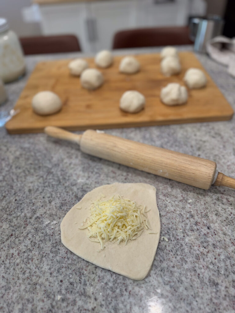 A piece of sourdough dough rolled out on a kitchen counter with a generous pile of shredded cheese in the center, ready to be shaped into a garlic cheese bomb. In the background, a wooden board holds several pre-shaped dough balls, and a rolling pin rests nearby, capturing the bread-making process in a cozy home kitchen.