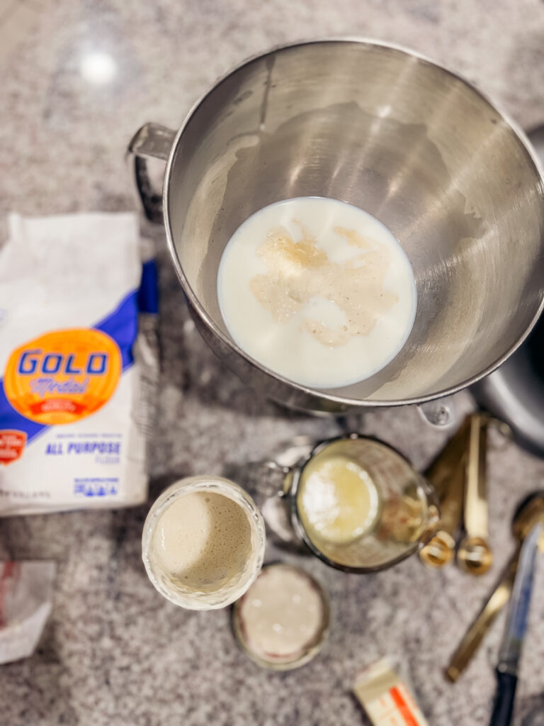 A top-down view of sourdough bread ingredients on a kitchen counter. A stand mixer bowl contains milk and sourdough starter. Nearby are a bag of all-purpose flour, a jar of active sourdough starter, melted butter in a glass measuring cup, measuring spoons, and a butter wrapper, all set up for making sourdough cheesy garlic bombs.