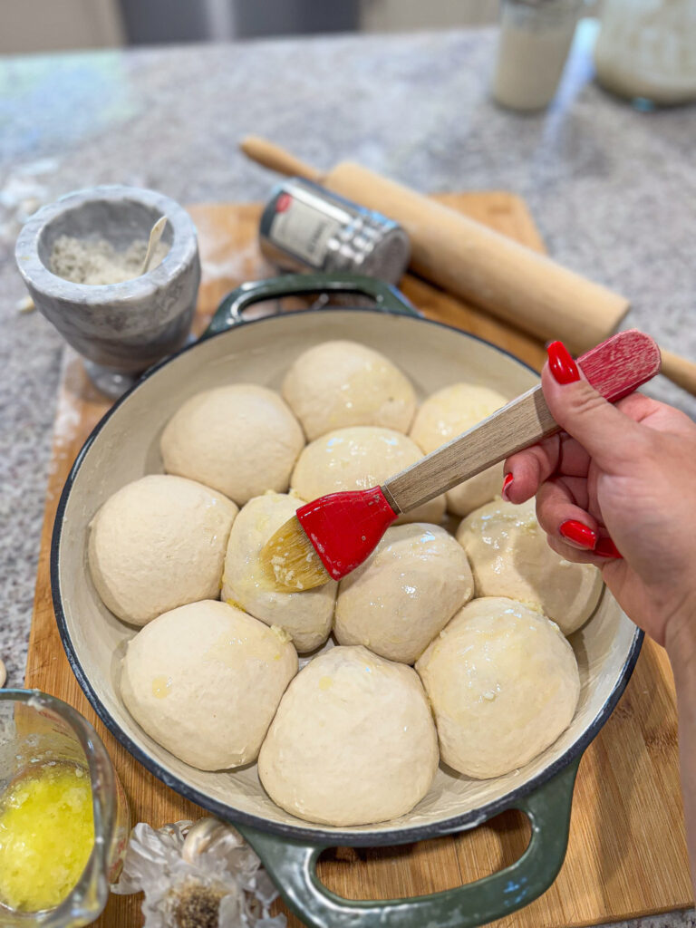 A hand uses a red pastry brush to apply melted garlic butter over unbaked sourdough garlic cheese bombs nestled in a round baking dish. The dough balls are smooth and plump, ready to rise and bake. Surrounding the dish are a mortar with minced garlic, a rolling pin, and a measuring cup of melted butter on a wooden board.