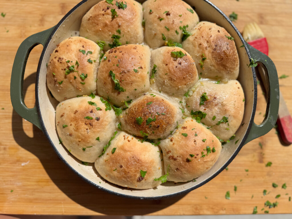 A fresh batch of golden-brown sourdough garlic cheese bombs baked in a round green Dutch oven. The rolls are puffed up and lightly crisp on top, sprinkled with minced garlic and fresh chopped parsley, glistening from a coating of garlic butter. A red pastry brush rests nearby on a wooden board.