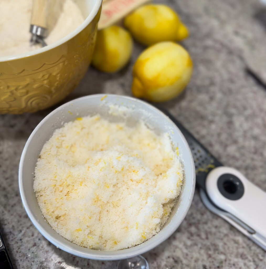 sourdough lemon bundt cake (2) A white bowl filled with lemon sugar, made by rubbing lemon zest into granulated sugar, sits on a kitchen counter next to whole lemons, a microplane zester, and a mixing bowl with flour.