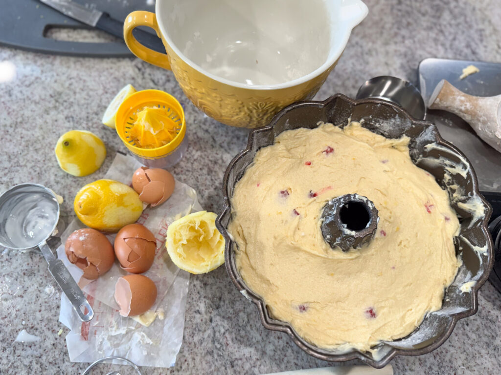 sourdough lemon bundt cake (5) A Bundt pan filled with lemon raspberry cake batter is ready to be baked, surrounded by baking ingredients including cracked eggshells, juiced lemons, a citrus juicer, measuring cups, and a large mixing bowl on a kitchen counter.