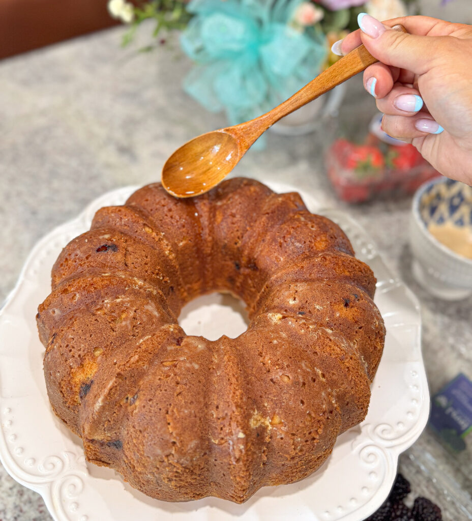 sourdough lemon bundt cake (6) A golden-brown sourdough lemon bundt cake sits on a white cake stand as a hand holds a wooden spoon, gently drizzling syrup over the warm cake, ready for the final glaze.