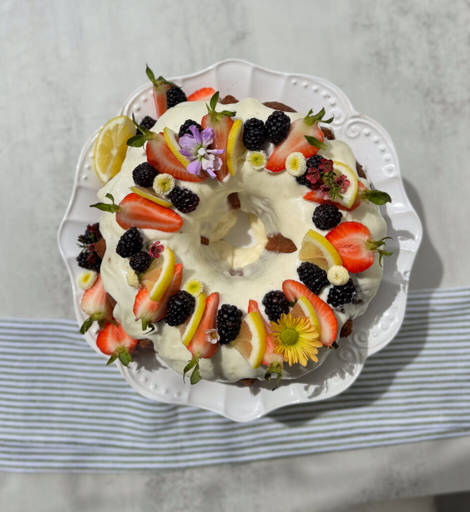 sourdough lemon bundt cake (7) A beautifully decorated sourdough lemon bundt cake topped with creamy white glaze, fresh strawberries, blackberries, lemon slices, and colorful edible flowers, displayed on a white scalloped cake stand with a striped cloth underneath.