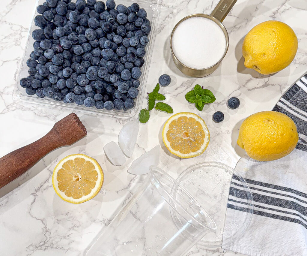 Overhead view of blueberry lemonade ingredients on a marble surface, including a container of fresh blueberries, two whole lemons, lemon halves, a measuring cup filled with sugar, fresh mint sprigs, ice cubes, a wooden muddler, a clear plastic cup with lid, and a striped kitchen towel.