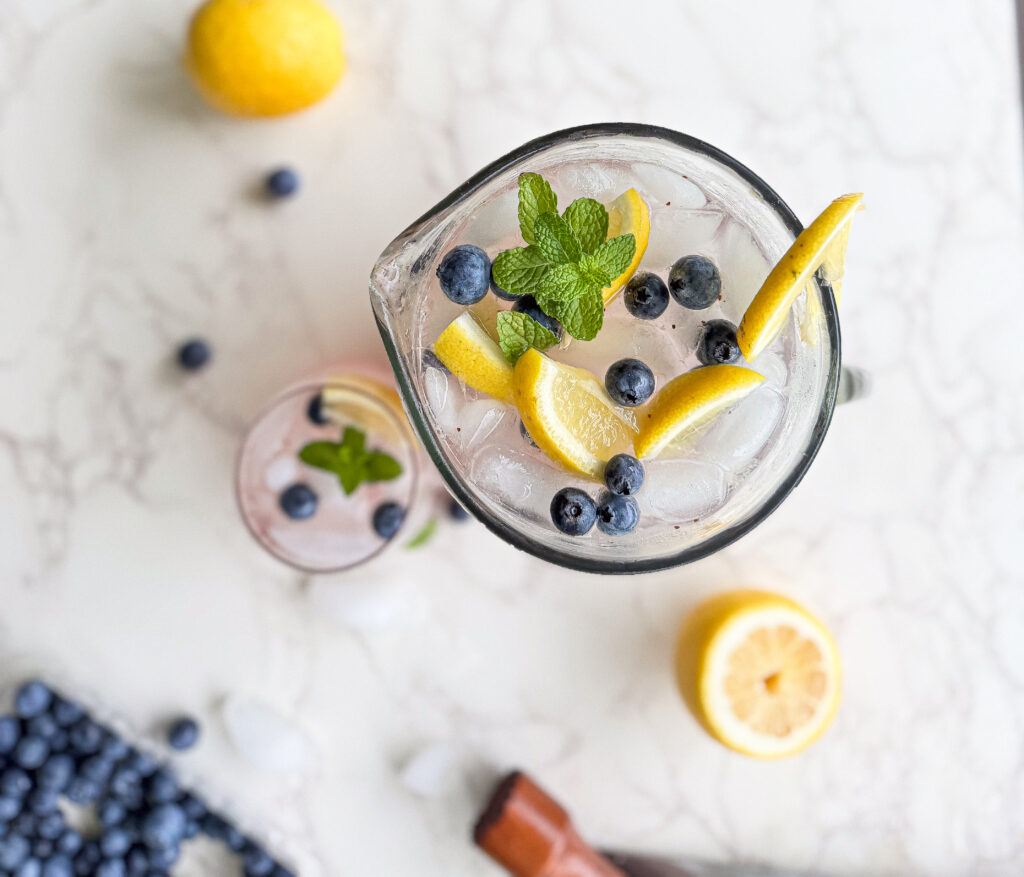 Top-down view of a pitcher and glass filled with iced blueberry lemonade, garnished with lemon slices, fresh blueberries, and mint leaves. The pitcher sits on a white marble surface surrounded by scattered blueberries, ice cubes, and a halved lemon.