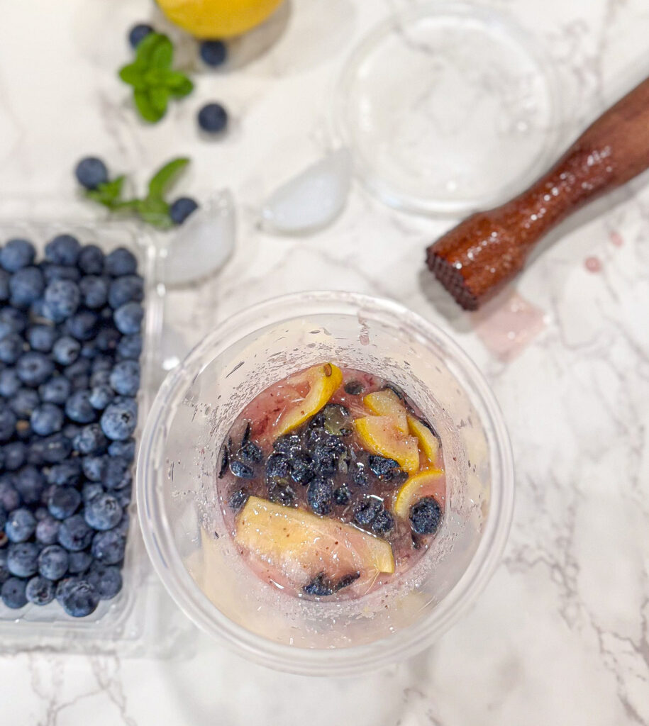 Top-down view of a plastic cup filled with muddled lemon slices and blueberries, surrounded by fresh blueberries in a container, a wooden muddler, ice cubes, and mint leaves on a white marble surface.