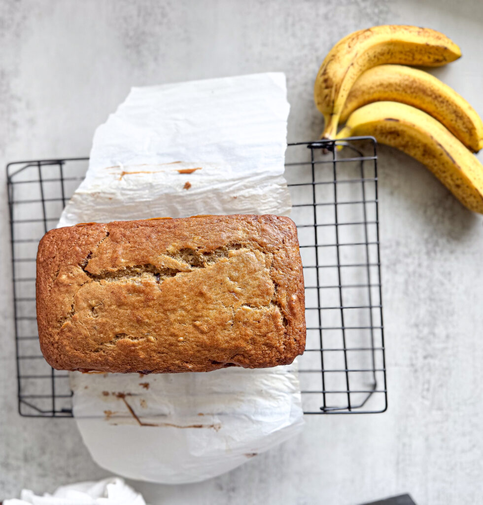 A freshly baked loaf of sourdough banana bread resting on a parchment-lined cooling rack, with ripe bananas in the background on a light gray surface.