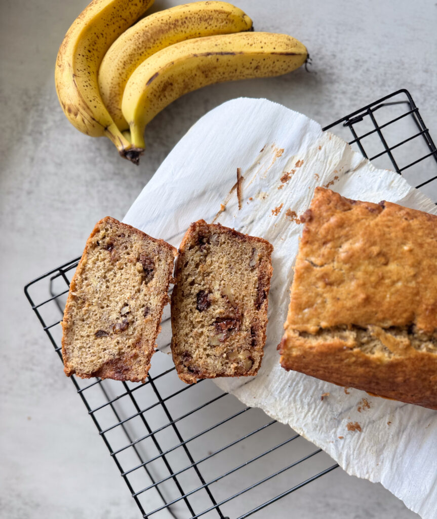 Two slices of sourdough banana bread with visible chocolate chips and walnuts on a cooling rack, next to the remaining unsliced loaf, with ripe bananas in the background.
