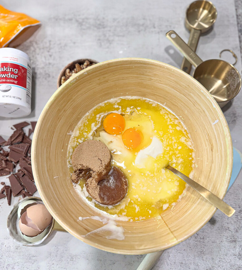 Mixing bowl filled with sourdough banana bread ingredients including two cracked eggs, brown sugar, melted butter, and yogurt, surrounded by baking items like chopped chocolate, measuring cups, baking powder, and walnut pieces on a light countertop.