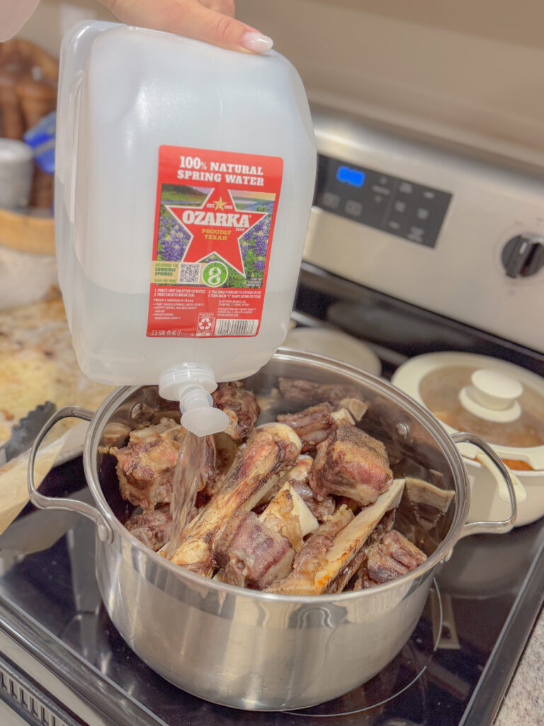 Water being poured from a large Ozarka spring water jug into a pot filled with roasted beef bones on the stovetop, beginning the process of simmering bone broth.