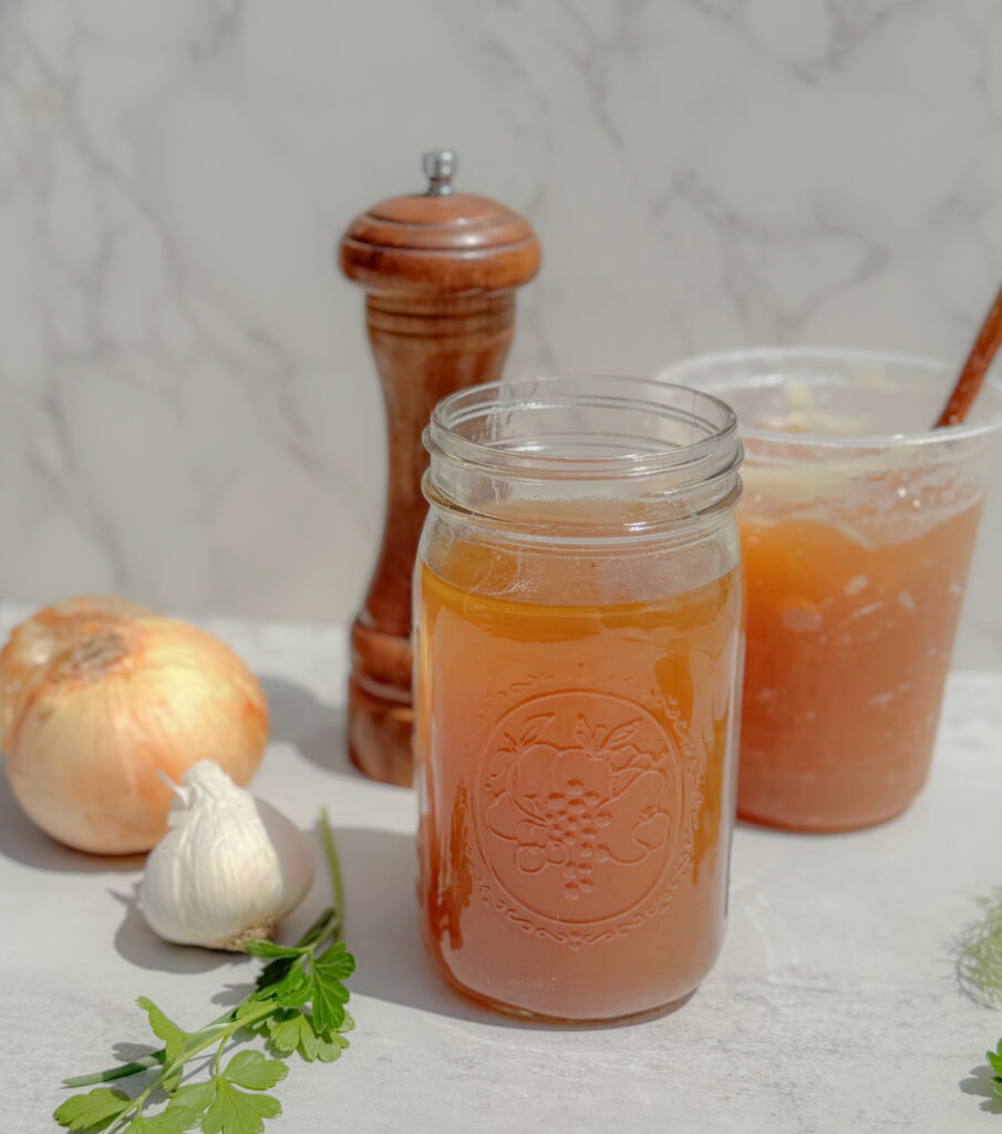 Close-up of a mason jar filled with rich golden beef bone broth, surrounded by a garlic bulb, yellow onion, fresh parsley, and a wooden pepper grinder, with a container of gelatinous broth in the background.