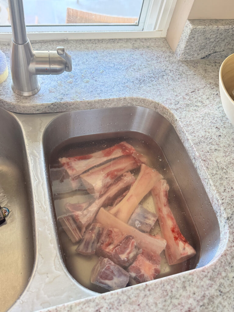 Raw beef bones soaking in cold water in a stainless steel kitchen sink to draw out impurities before roasting.