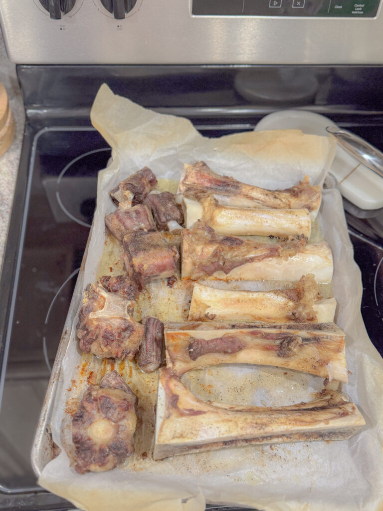 Roasted beef bones, including marrow bones and oxtail, laid out on a parchment-lined baking sheet fresh out of the oven, ready for broth making.