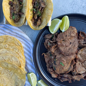 Two beef tacos in holders, a black plate with cooked beef topped with herbs, a stack of corn tortillas, and lime wedges on a gray surface.