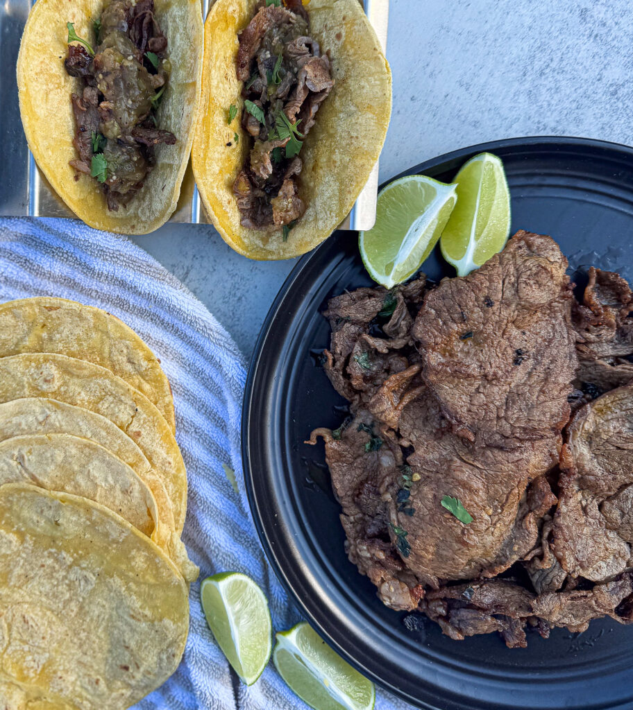 Grilled carne asada served on a black plate with lime wedges, next to tacos filled with steak and salsa. Fresh corn tortillas and a striped towel complete the setup.