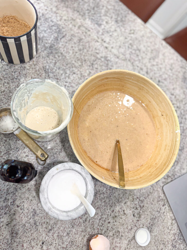 Overhead view of a kitchen counter with ingredients being prepped for baking sourdough banana cake, including a large bowl of wet batter, a glass jar of sourdough starter, a container of sugar with a spoon, vanilla extract bottle, a cracked egg shell, and measuring spoons.