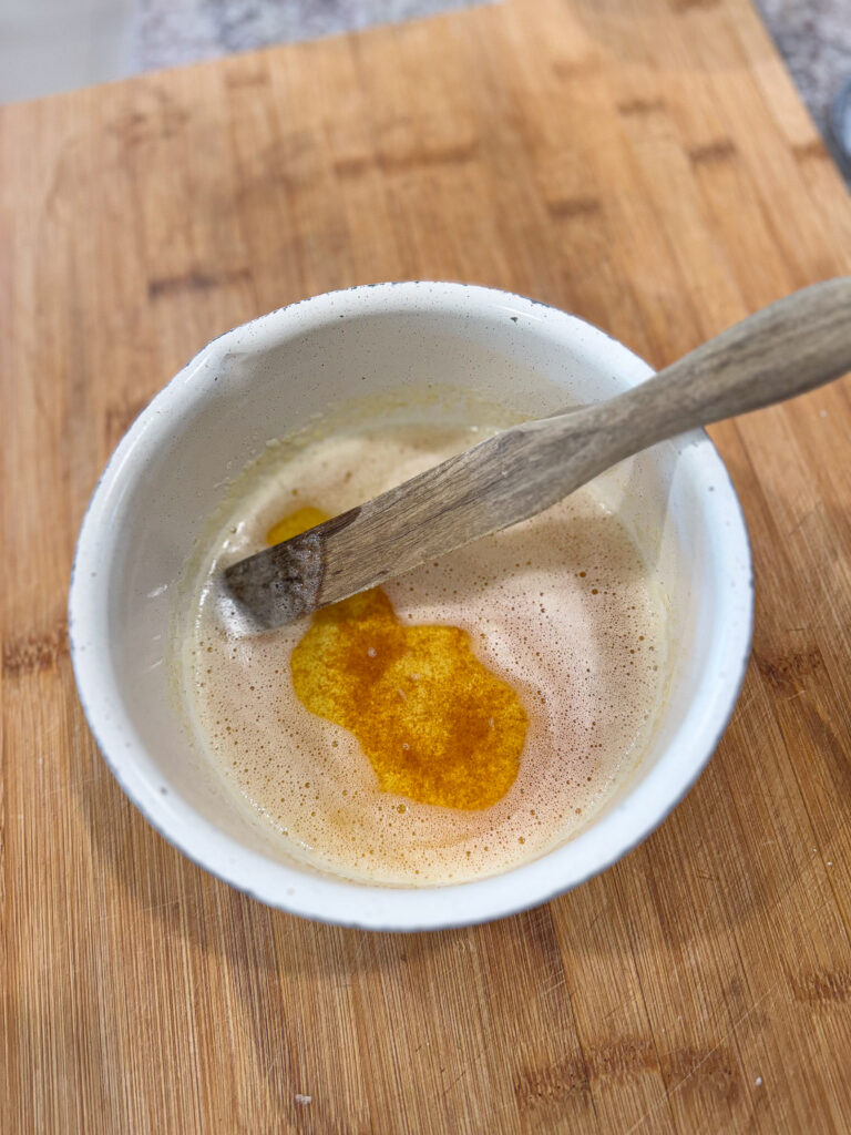 Close-up of a small white bowl filled with freshly browned butter, with golden amber specks and foamy edges, resting on a wooden cutting board with a wooden spatula inside.