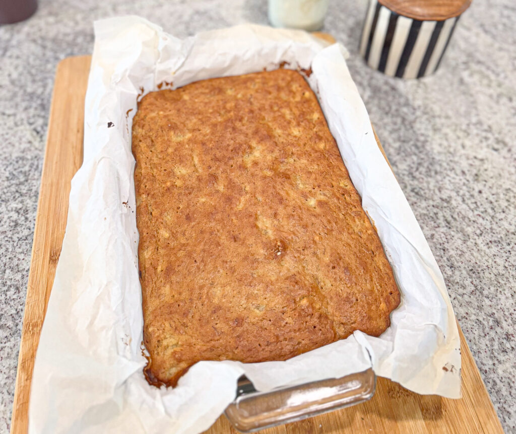 Freshly baked sourdough banana cake in a parchment-lined glass loaf pan, resting on a wooden cutting board with a golden-brown top and moist texture, cooling on a kitchen counter.