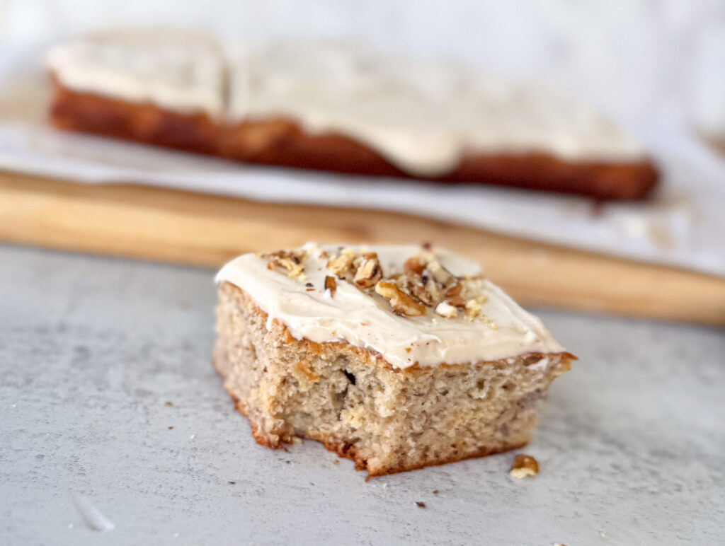 Close-up of a single slice of sourdough banana cake topped with whipped brown butter cream cheese frosting and chopped walnuts, with the rest of the frosted cake blurred in the background on a wooden board